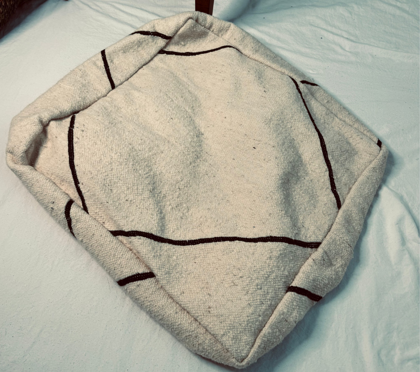 A square off-white wool pet bed with dark brown stitched geometric lines, placed on a white sheet with a wooden stool leg visible in the background.