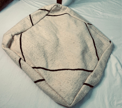 A square off-white wool pet bed with dark brown stitched geometric lines, placed on a white sheet with a wooden stool leg visible in the background.