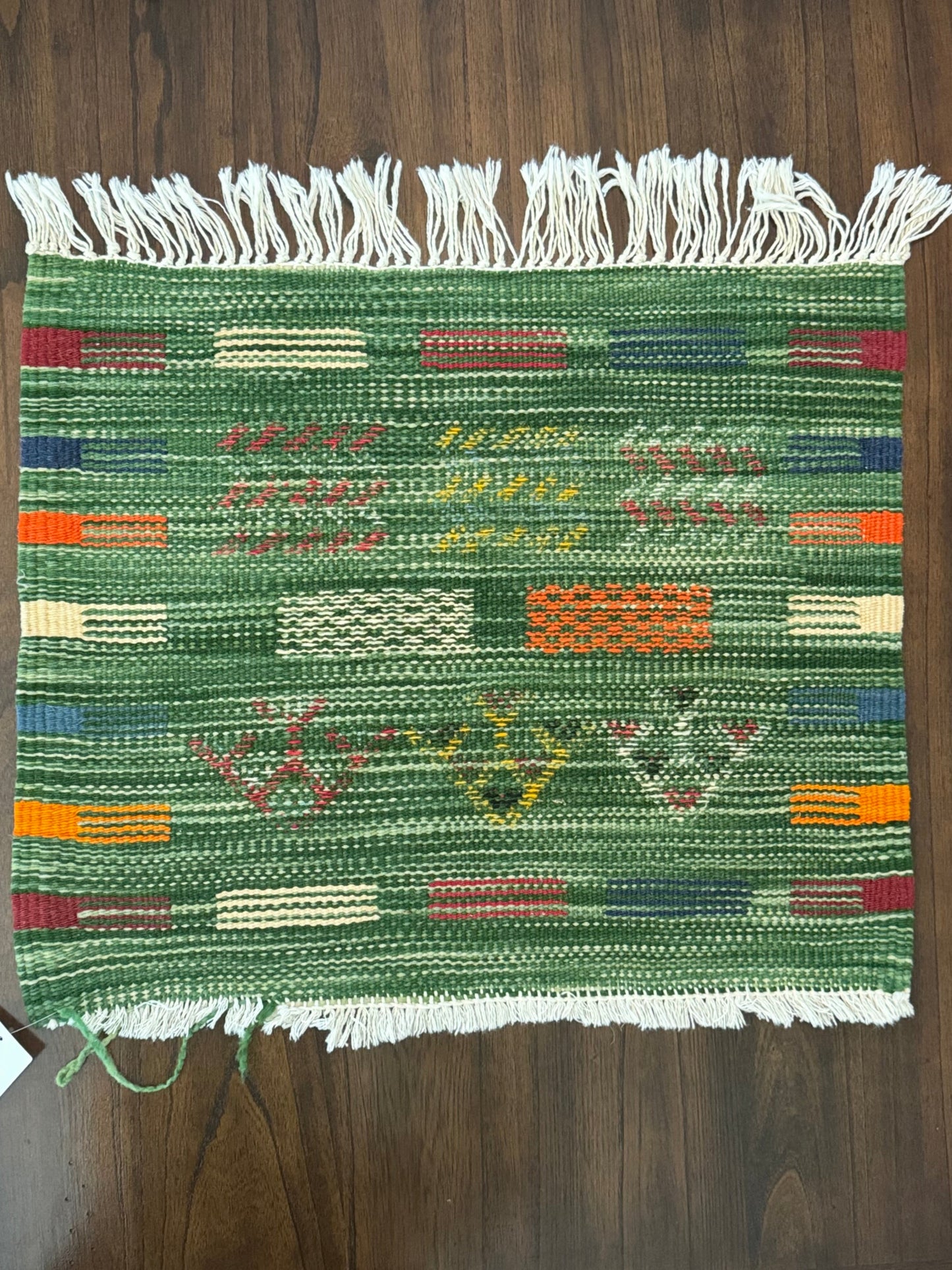 A woven green textile rug or throw with multicolored geometric stripes and white fringe on both short ends, laid flat on a dark wood floor.