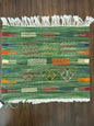 A woven green textile rug or throw with multicolored geometric stripes and white fringe on both short ends, laid flat on a dark wood floor.